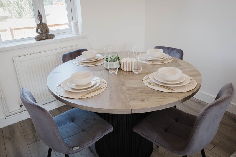 Round wooden dining table set with place settings and upholstered chairs, window in background
