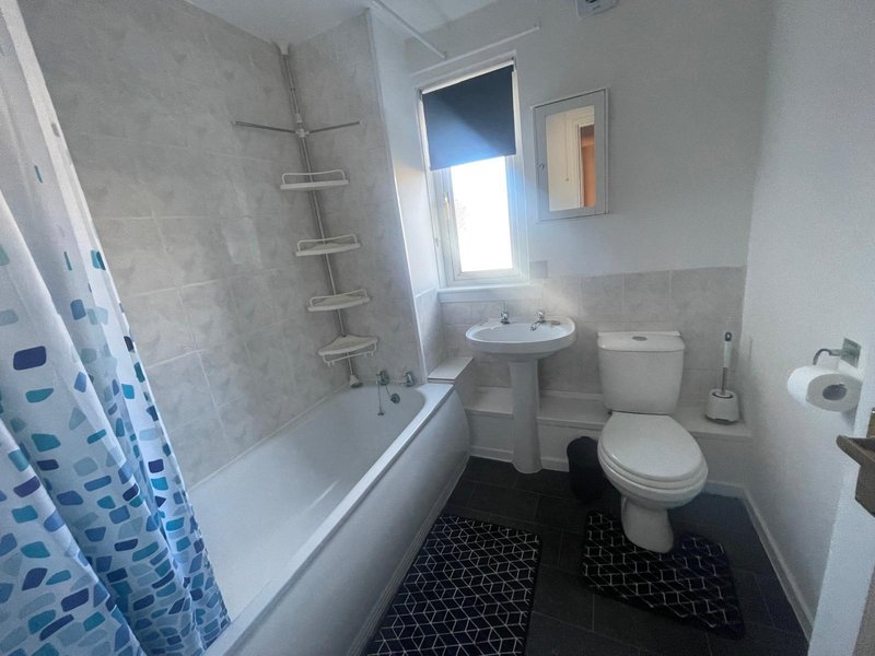 Small bathroom with bathtub, pedestal sink, toilet, and window with blue shower curtain