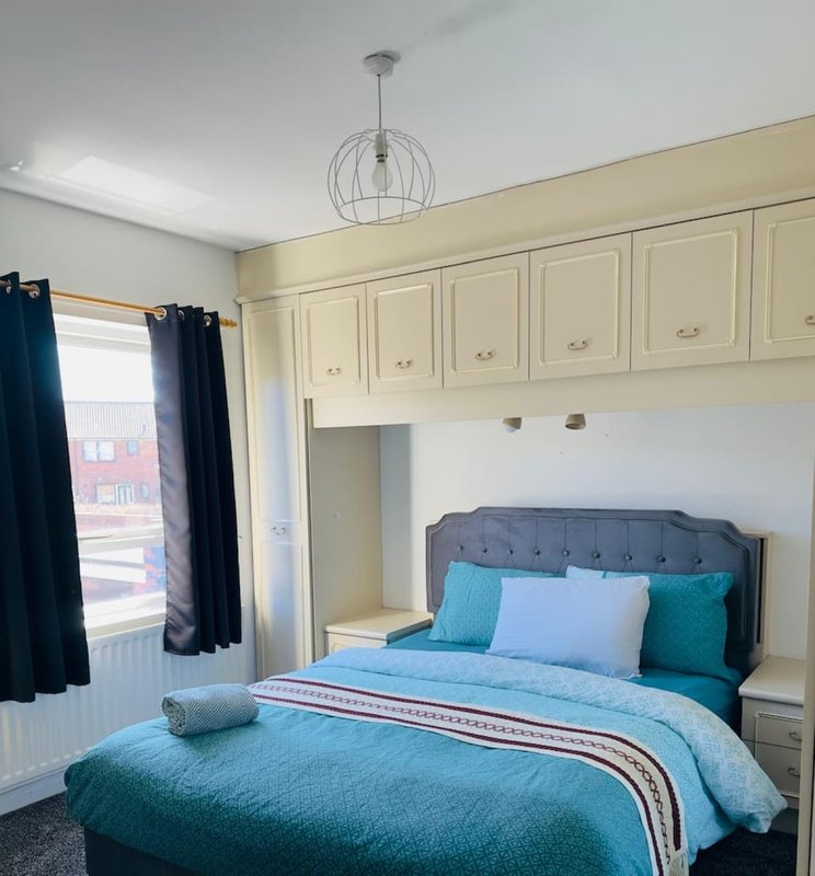 Bedroom with double bed, turquoise bedding, built-in cream wardrobes, pendant light, and window with dark curtains