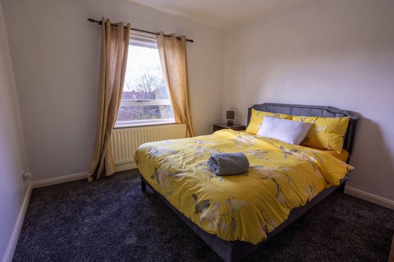 Bedroom with double bed, yellow bedding, dark carpet, window with tan curtains