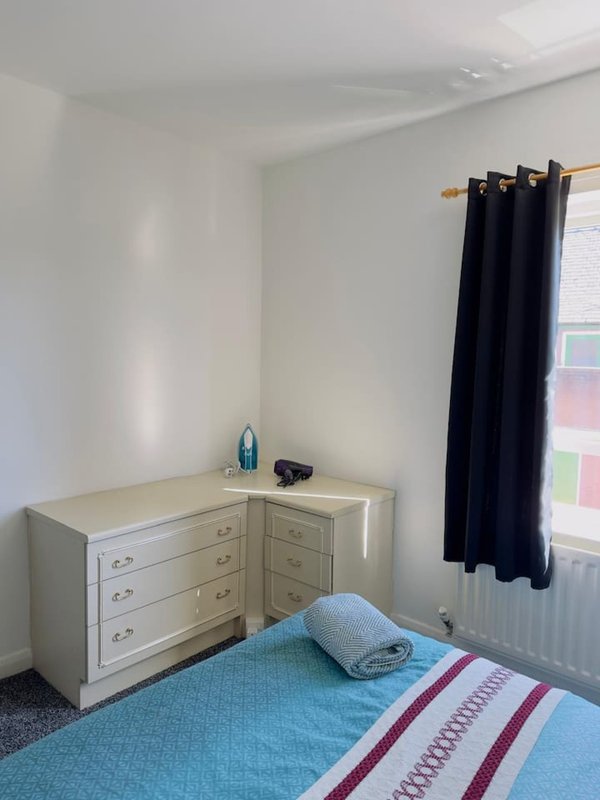 Bright bedroom with double bed, wooden dresser, window with black curtains and radiator