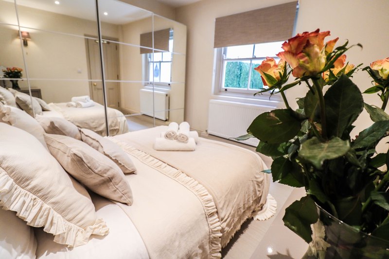 Bright bedroom with double bed, multiple pillows, ensuite bathroom visible through glass door, fresh flowers on nightstand