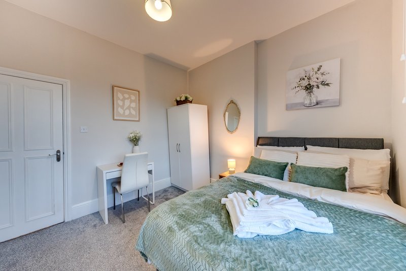 Bedroom with double bed, green bedding, white wardrobe, desk and framed wall art