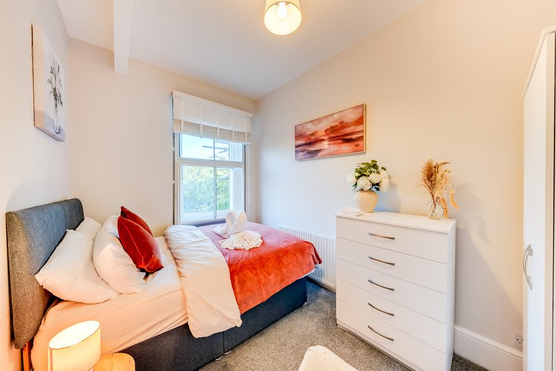 Bright bedroom with double bed, red throw, white chest of drawers and window with natural light