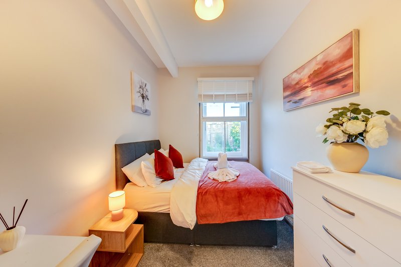 Bright bedroom with double bed, red throw, bedside lamp, dresser, and window view