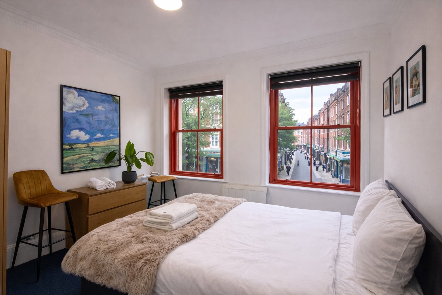 Bright bedroom with double bed, wooden desk, chair, framed artwork and large windows with red frames