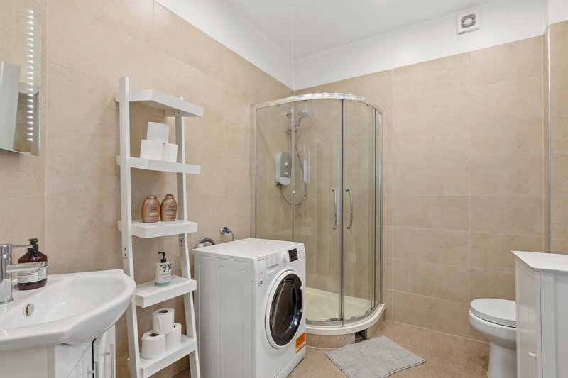 Beige bathroom with shower enclosure, washing machine, sink, toilet and white shelving