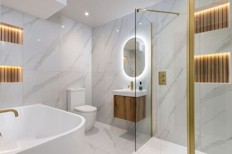 Modern bathroom with freestanding tub, marble walls, illuminated mirror and gold fixtures