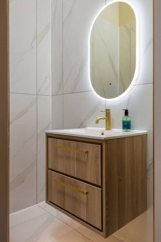 Modern bathroom with illuminated oval mirror, wooden vanity, white sink and marble-effect wall tiles