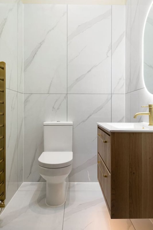 Modern bathroom with white marble tiles, toilet, vanity with wooden cabinetry and brass fixtures