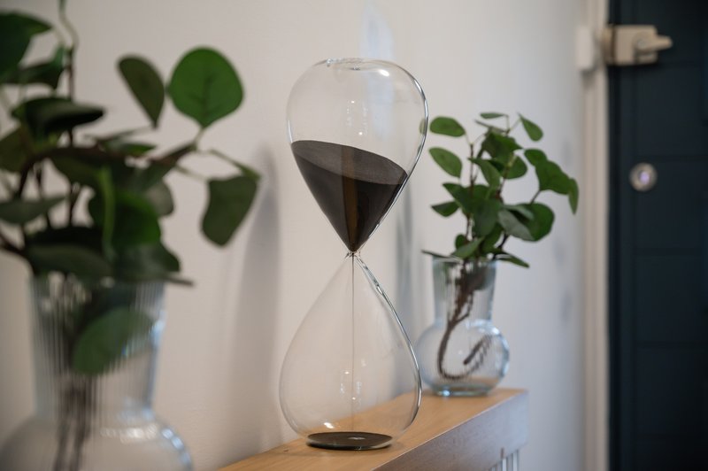 Hourglass with black sand on wooden shelf surrounded by potted green plants