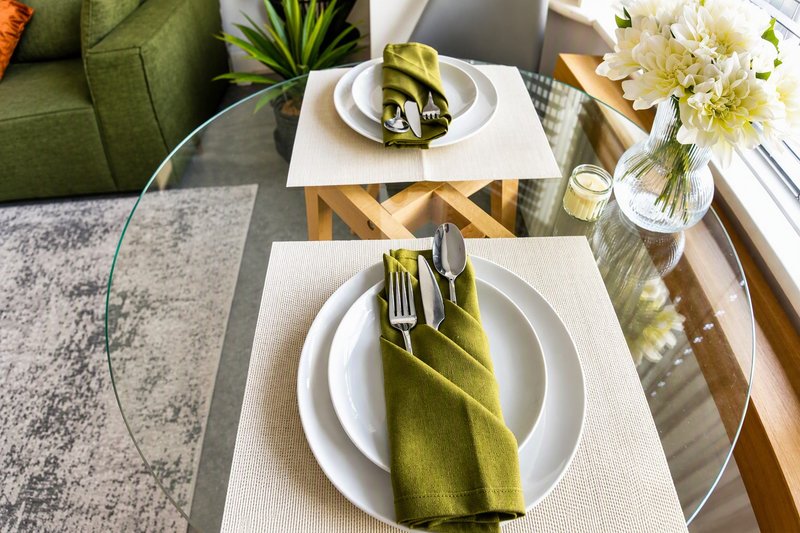 Dining table set for two with white plates, green napkins, flowers in vase and green sofa