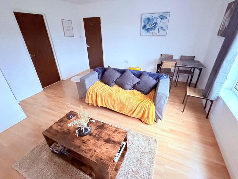 Open-plan living room with wooden flooring, sofa with yellow and purple bedding, rustic coffee table, and dining area
