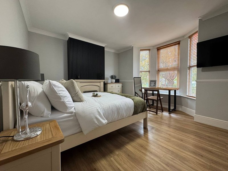 Modern bedroom with white bedding, wooden floor, desk area and large windows with blinds
