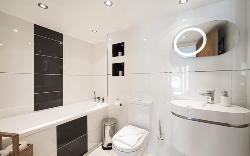 Modern bathroom with white tiles, black accent panels, curved sink, illuminated mirror and bathtub