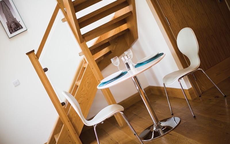 Compact dining area with wooden staircase, small table, and white chairs on wooden floor