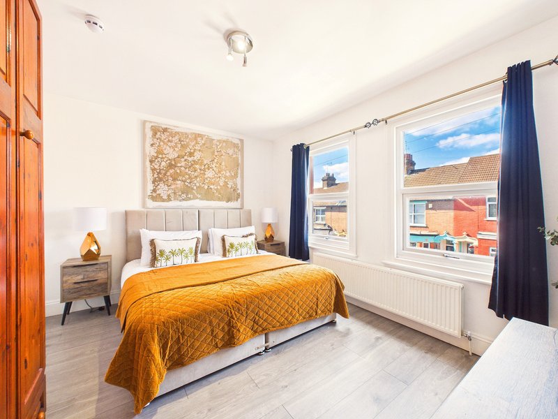 Bright bedroom with double bed, golden bedspread, wooden wardrobe and large windows overlooking street