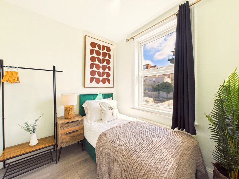 Bright bedroom with single bed, green headboard, wooden nightstand, and window with black curtains