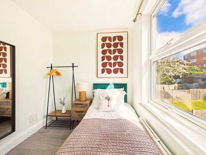 Bright bedroom with single bed, wooden nightstand, floor lamp, and large windows overlooking garden