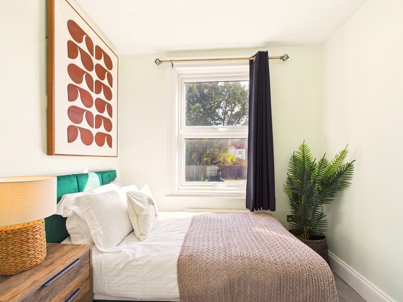 Bright bedroom with double bed, white bedding, window with black curtains, wall art and potted plant