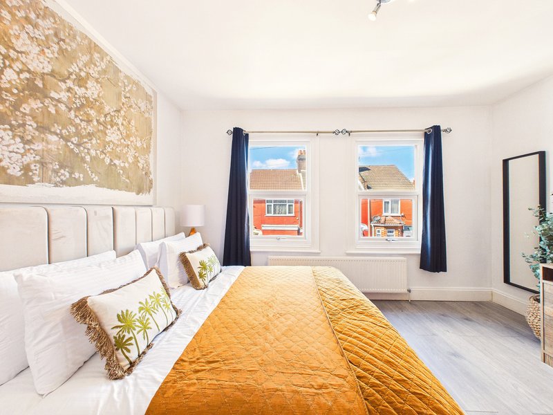 Bright bedroom with white panelled bed, yellow bedding, decorative pillows, and windows overlooking residential street