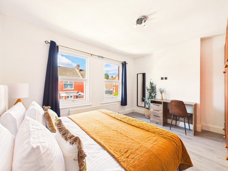 Bright bedroom with double bed, golden throw, desk, chair, and large windows overlooking street