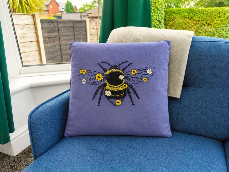 Blue upholstered sofa with purple cushion featuring bee embroidery, window view to garden