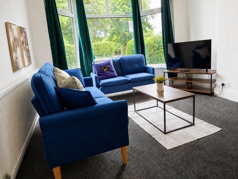 Bright living room with blue sofas, coffee table, TV, and large windows overlooking garden