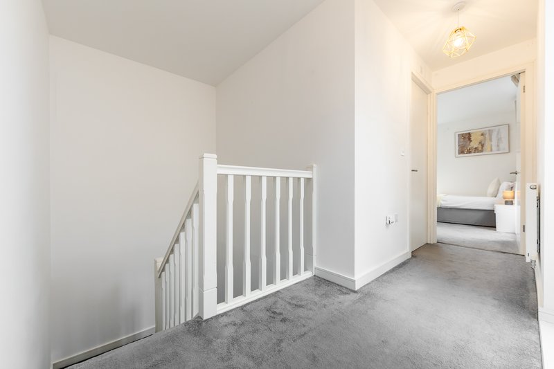 Bright hallway with white stair railing, grey carpet, and open doorway to bedroom