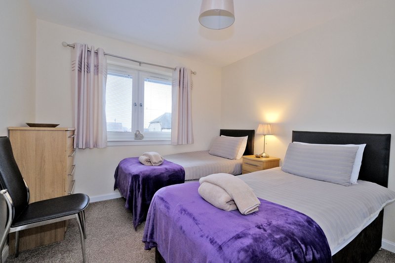 Bright bedroom with two single beds, purple throws, wooden chair and large window with curtains