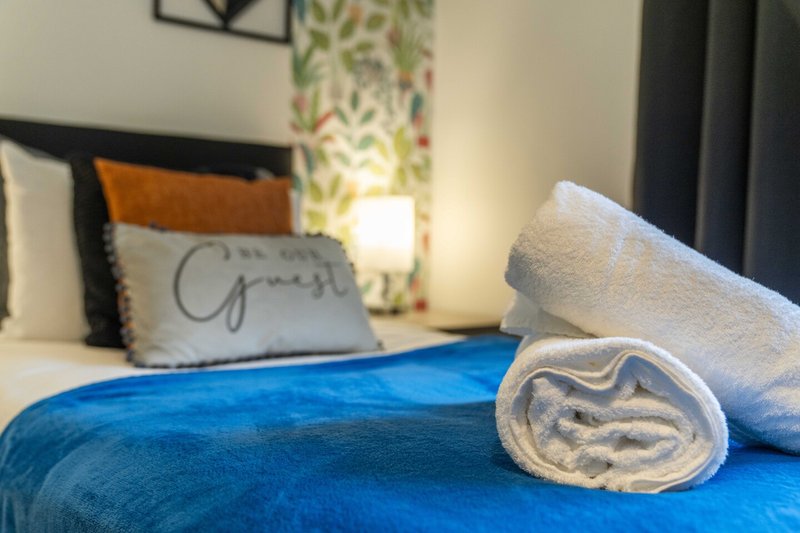 Bedroom with blue bedspread, rolled towels, decorative pillow, and bedside lamp