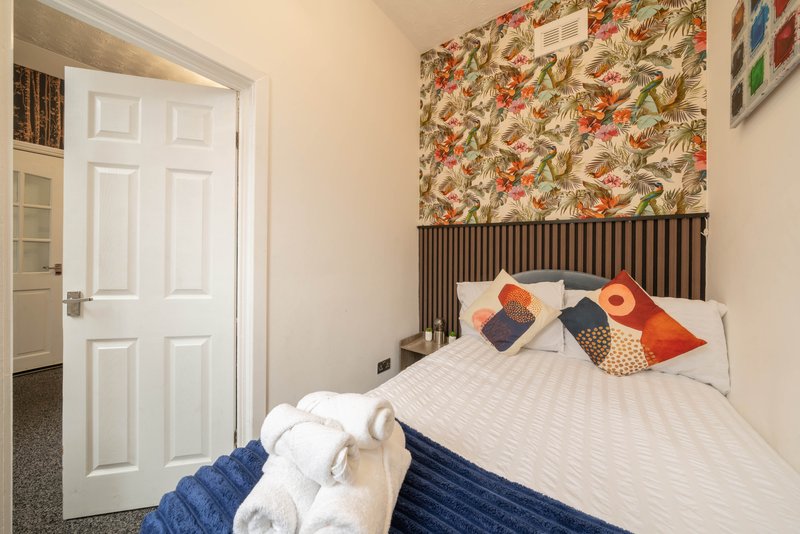 Bedroom with double bed, colorful cushions, floral wallpaper headboard, white door and cream walls