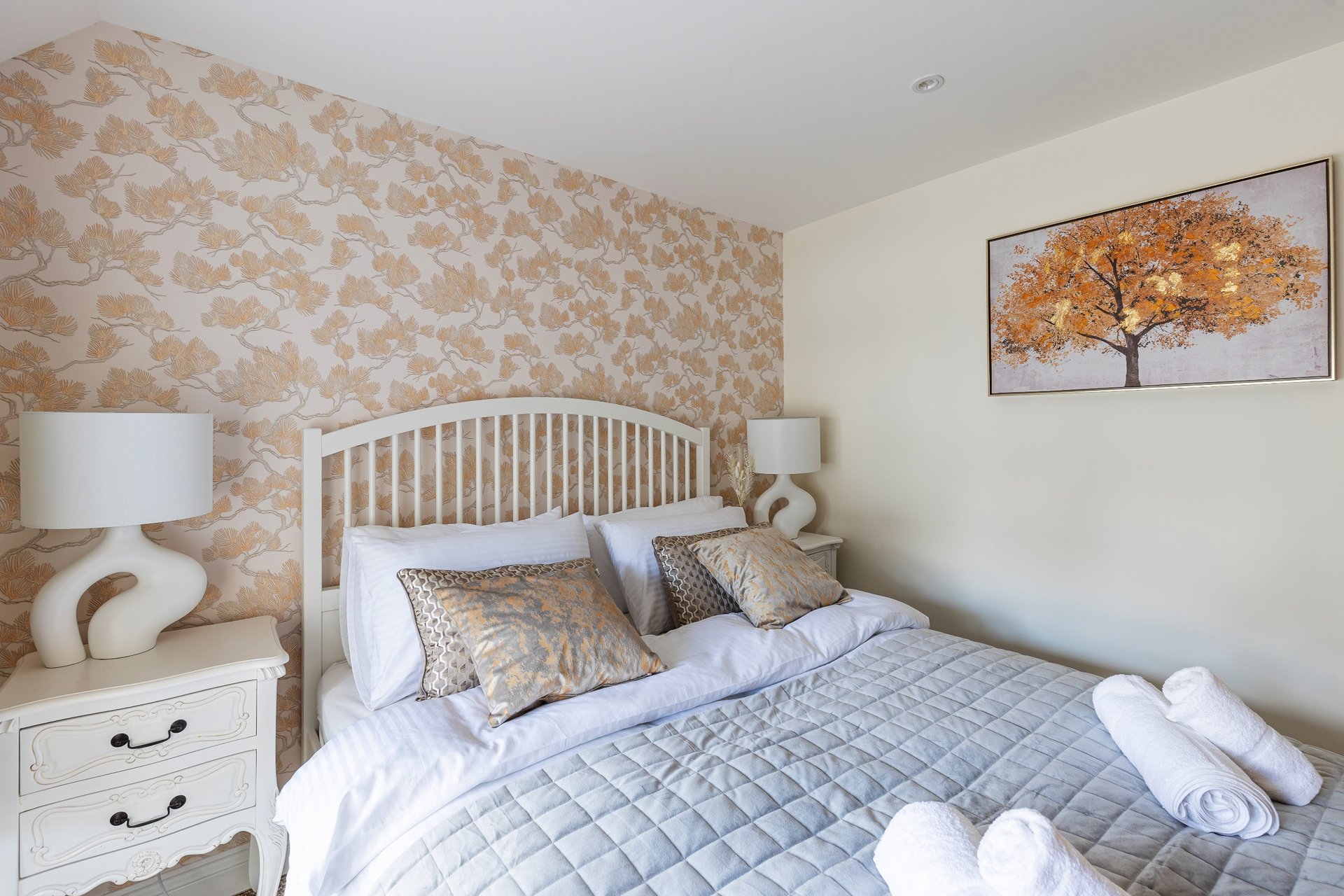 Bedroom with white wooden bed frame, patterned wallpaper, bedside table and framed wall art