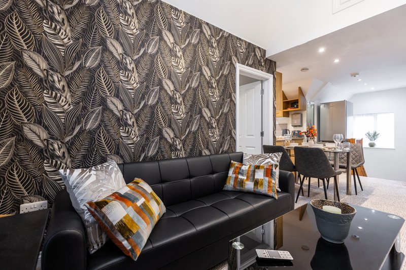 Modern living room with black leather sofa, patterned feature wall, and open-plan kitchen area