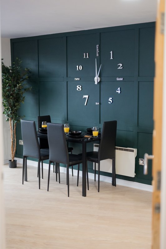 Dining area with black table, chairs, and large wall clock against dark teal panelled wall