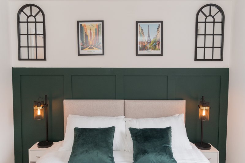 Bedroom with green upholstered headboard, white bedding, dark green velvet pillows and wall sconces