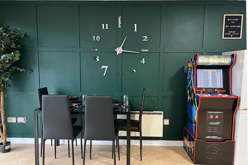 Dining area with green panelled wall, wall clock, dining table, chairs, and arcade machine