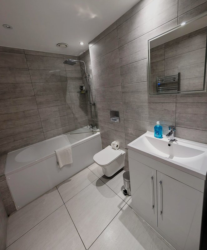 Modern bathroom with gray tile walls, white bathtub, toilet, and vanity sink with mirror