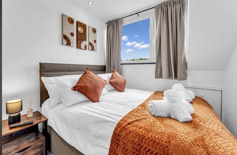Bright bedroom with double bed, orange throw, white pillows, wooden nightstand and window with countryside view
