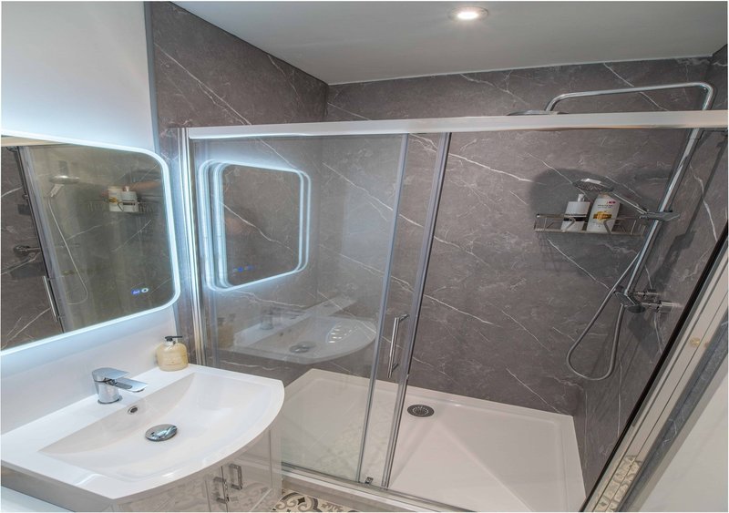 Modern bathroom with gray tile walls, white sink, illuminated mirror, and walk-in shower enclosure