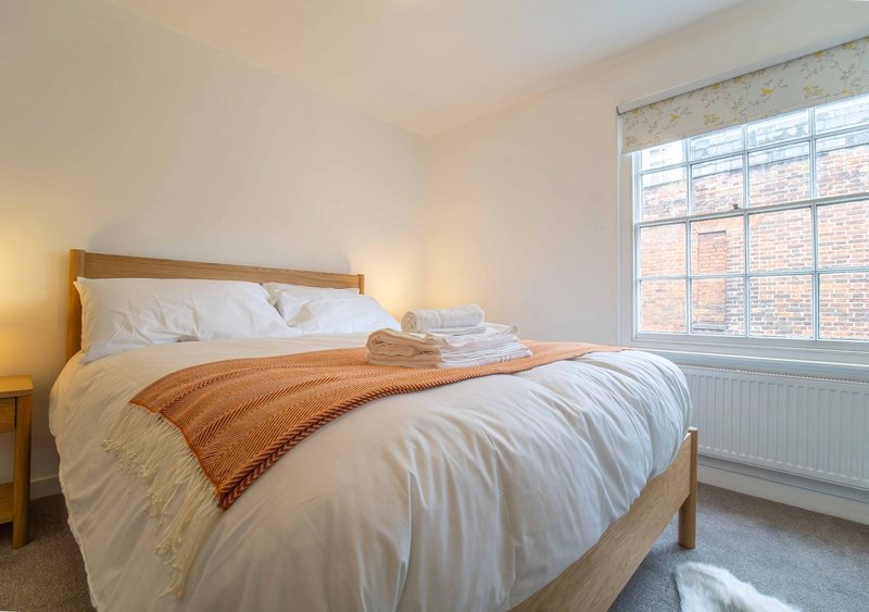 Bright bedroom with wooden bed frame, white bedding, orange throw blanket, and large window overlooking brick building