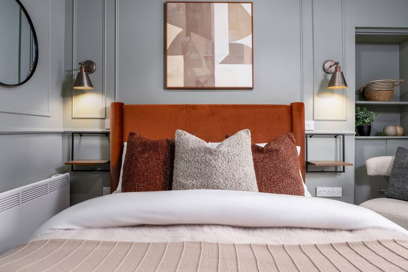 Modern bedroom with orange upholstered headboard, white pillows, rust and cream cushions, wall sconces and framed artwork
