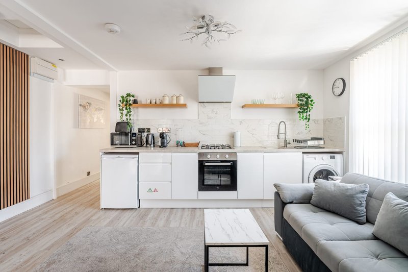 Open-plan kitchen and living room with white cabinetry, modern appliances, wooden shelving, and gray sofa