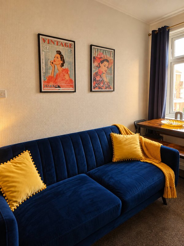 Living room with blue velvet sofa, yellow cushions, vintage wall art, and window with curtains