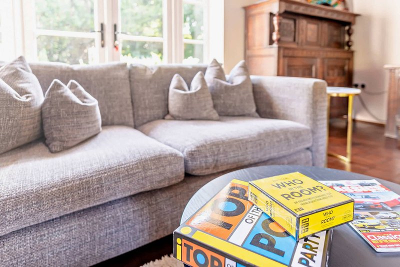 Spacious living room with gray upholstered sofa, cushions, board games on table, wooden furniture and large windows