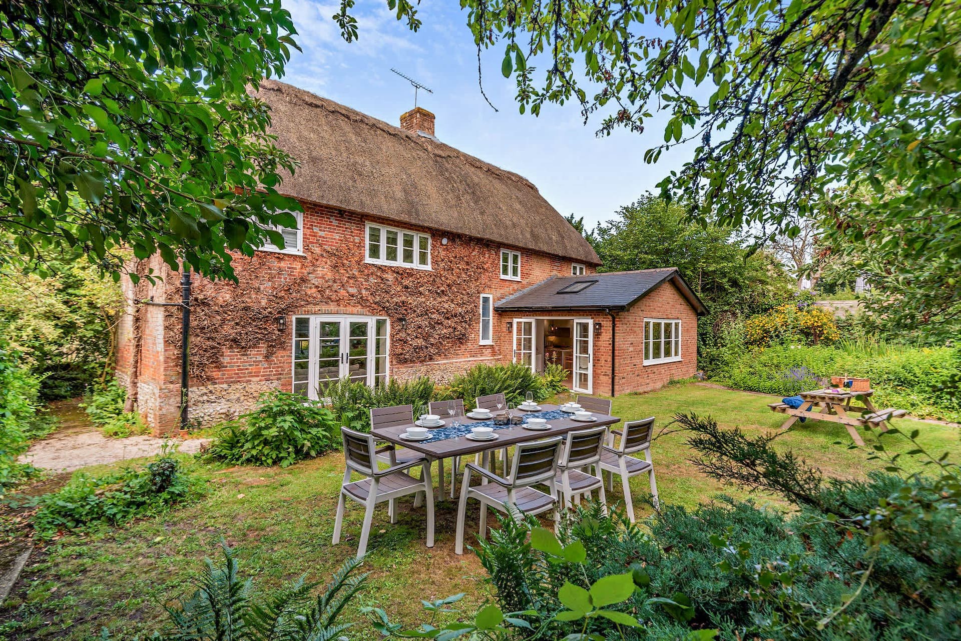 Red brick cottage with thatched roof, garden table and chairs on lawn
