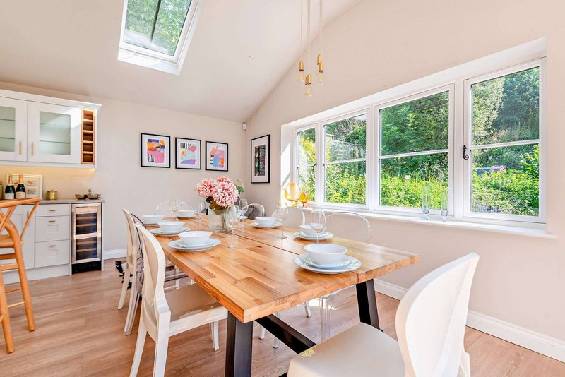 Bright dining room with wooden table, white chairs, pink flowers, and large windows overlooking garden