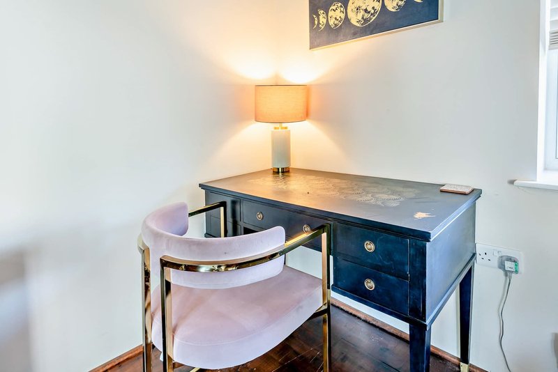 Compact home office with dark blue desk, pink upholstered chair, table lamp and wall art