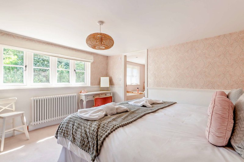 Bright bedroom with double bed, patterned bedding, pink cushion, wicker ceiling light and large windows