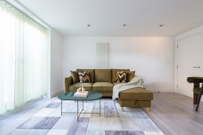 Modern living room with olive-green sectional sofa, glass coffee table, geometric area rug, and large windows with white curtains